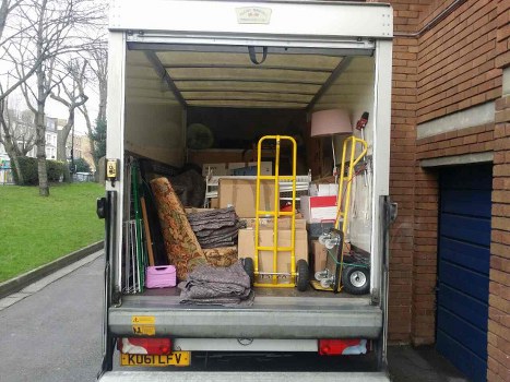 The image shows the open cargo area of a white waste collection van parked on a driveway next to a brick building. Inside the van, there are various items including floral-patterned upholstered chairs, some with fabric coverings, stacked and leaning against the sides. There is a yellow hand truck positioned vertically near the center, along with a chrome-colored shopping trolley, and a small push lawn mower with a black handle and grey body. Also visible are cardboard boxes, a pink plastic container, and a table lamp with a beige shade. The interior ceiling of the van is exposed timber with some hanging hooks. The van is parked in an area that appears to be on a residential street, with a trimmed lawn, trees, and other houses visible in the background, suggesting a typical London suburb. Business Waste Removal Stoke Newington offers reliable rubbish removal services, and this image illustrates a collection of household items ready for disposal or recycling, consistent with their local rubbish clearance offerings in Stoke Newington, N16.