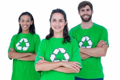 A group of three individuals standing outdoors, each wearing bright green T-shirts featuring a white recycling symbol on the front. The person in the center is a woman with dark curly hair, smiling directly at the camera with her arms crossed. To her left is a woman with straight black hair, also smiling with arms crossed, and to her right is a man with short dark hair and a beard, smiling and standing with his arms crossed as well. The background appears to be an outdoor environment, possibly a park or urban area. The bright green T-shirts and recycling symbols suggest their involvement in waste management or environmental initiatives, aligning with the services offered by businesses such as rubbish removal or waste disposal in the Stoke Newington area. The image conveys enthusiasm and teamwork within a professional context focused on sustainable waste solutions.