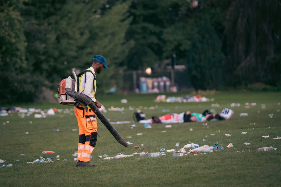 A sanitation worker wearing a blue cap, high-visibility orange trousers with reflective stripes, and a vest operates a leaf blower to clear away litter from a grassy park area. The worker is positione