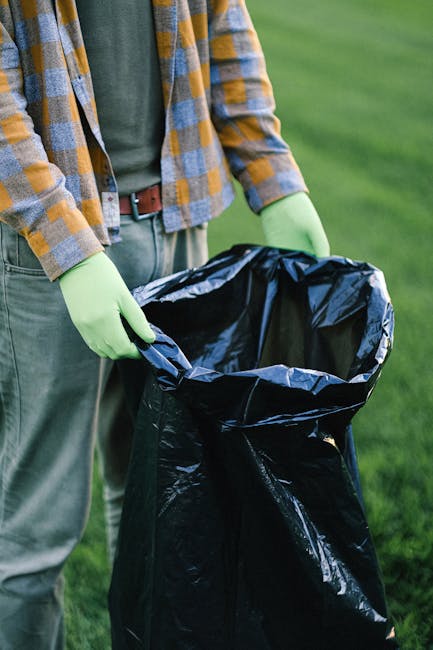 A sanitation worker wearing a blue cap, high-visibility orange trousers with reflective stripes, and a vest operates a leaf blower to clear away litter from a grassy park area. The worker is positione